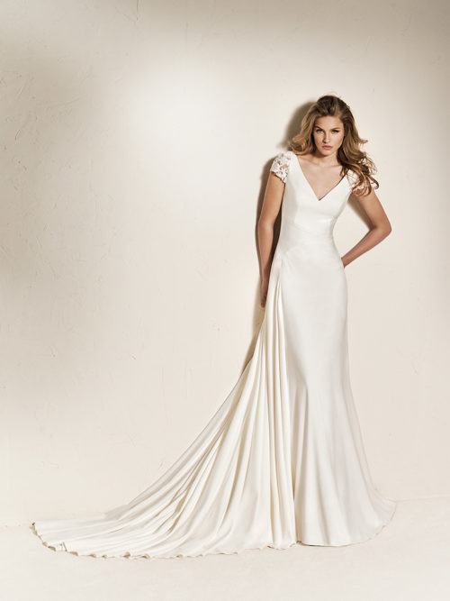 The simplicity of the crepe is what gives form and life to this spectacular flared wedding dress with cap sleeves in lace. A beautiful design that combines a fitted bodice featuring a V-neck in the front and back with a luxurious skirt and long train. A wedding dress with beaded appliqués on the sleeves that tiny sparkles to the final look.